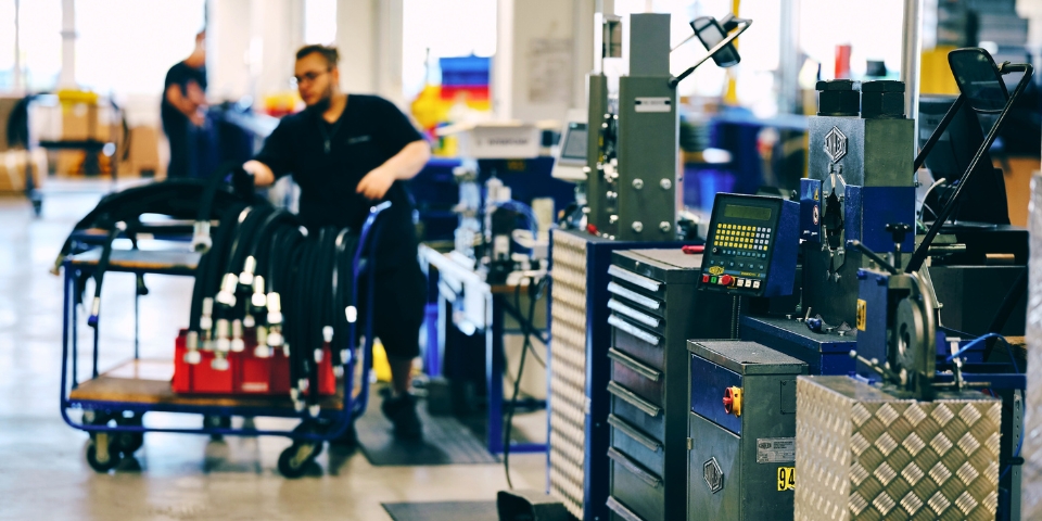 Ein Arbeiter in einer Fabrik bedient Maschinen zur Fertigung von Hydraulikschläuchen und bewegt diese  auf einem Wagen. Der Arbeitsplatz ist voll mit Industriegeräten, Schubladen und Monitoren. Die Atmosphäre ist geschäftig und gut organisiert.