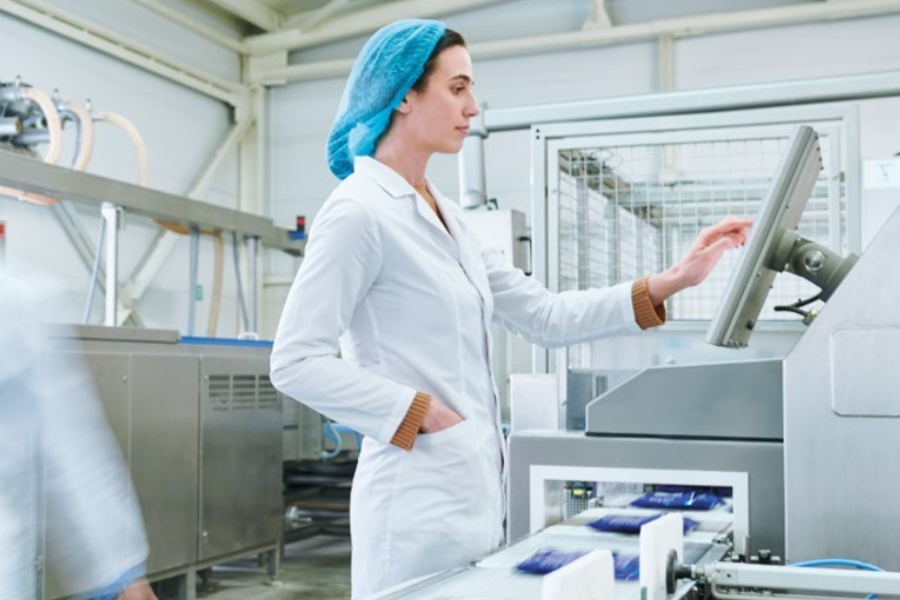 Eine Person im weißen Laborkittel und mit blauem Haarnetz bedient einen Touchscreen an einer Industrieanlage. In der Nähe transportieren Förderbänder verpackte Produkte in dieser sauberen, organisierten Fabrikumgebung.