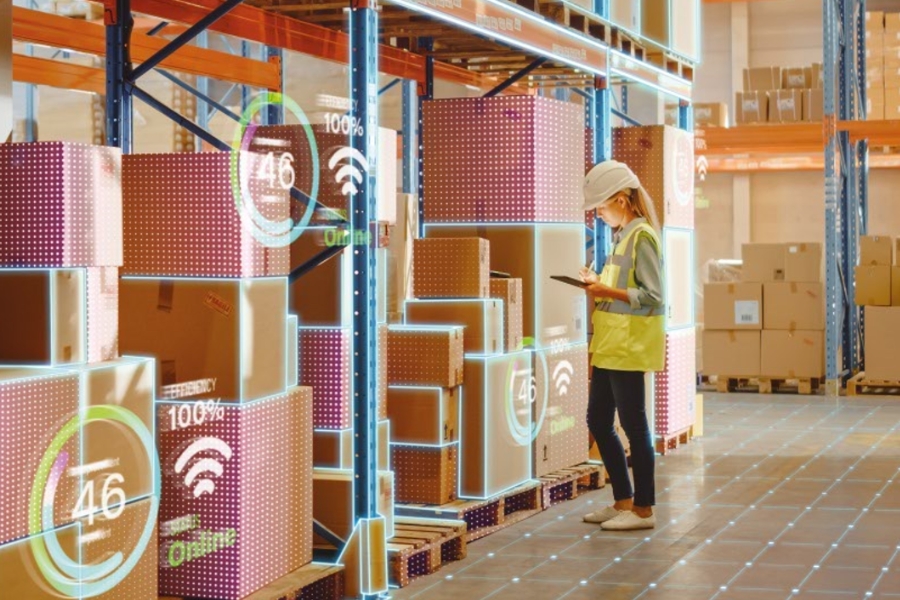 Eine Arbeiterin mit Schutzhelm und Sicherheitsweste nutzt ein Tablet in einem Lager. Die Regale sind randvoll mit Kartons, digitale Overlays zeigen Symbole und Texte wie „100 %“ und „46 Online“ und präsentieren damit die vernetzte Technologie.