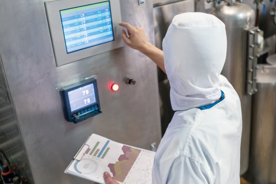 Eine Person in Schutzkleidung bedient ein Bedienfeld in einem Industrieumgebung. Sie hält ein Klemmbrett mit Grafiken und Diagrammen in der Hand und überwacht die auf Bildschirmen angezeigten Daten. Im Hintergrund sind Metalltanks deutlich zu erkennen.