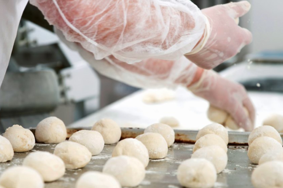 Hände in Schutzhandschuhen formen Teigbällchen auf einem bemehlten Backblech.